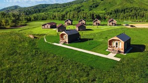 A series of small, uniform cabins with a central walkway, surrounded by lush green fields and a forested hill in the background. - Park-Hotel Zarechye  (Bystrukha)