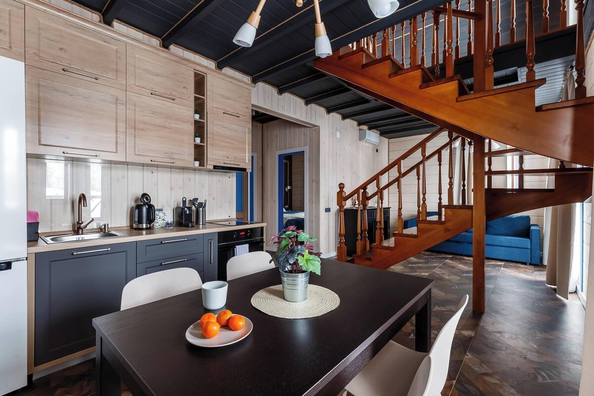 Una cocina moderna con armarios oscuros, una escalera de madera y un comedor con una mesa preparada para dos.