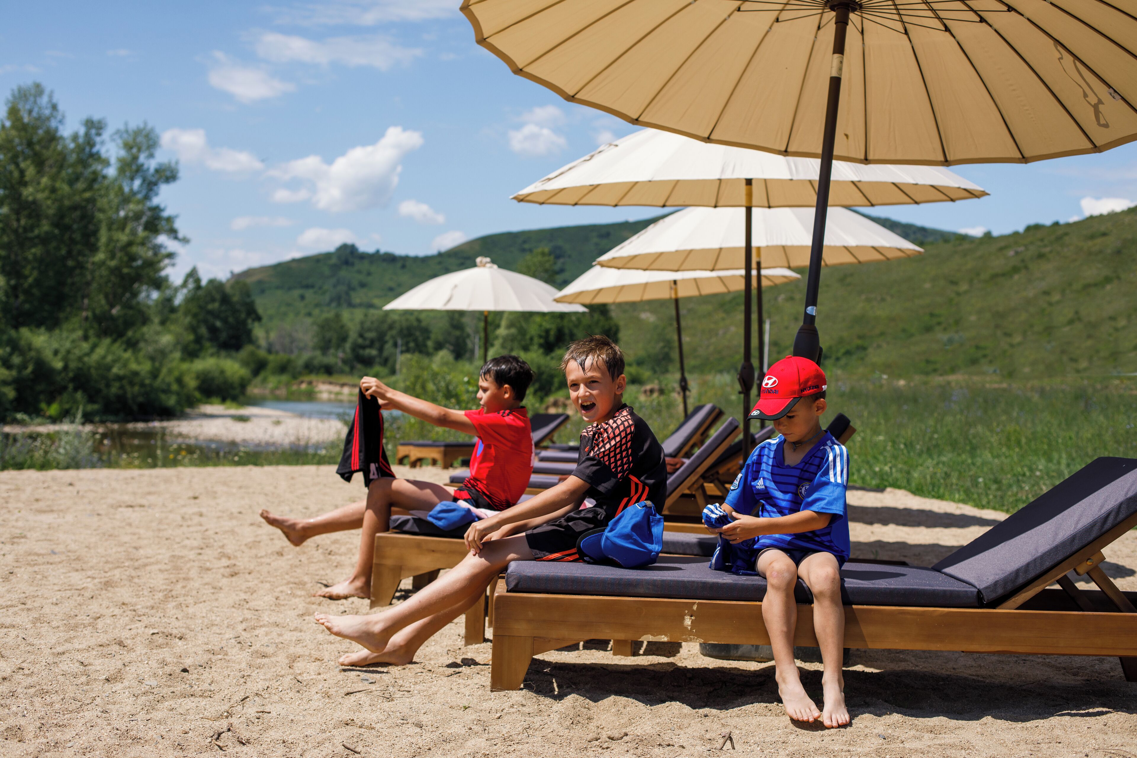Trois enfants sont assis sur des transats, à l’ombre de parasols, sur une plage de sable, avec un paysage vallonné en arrière-plan.