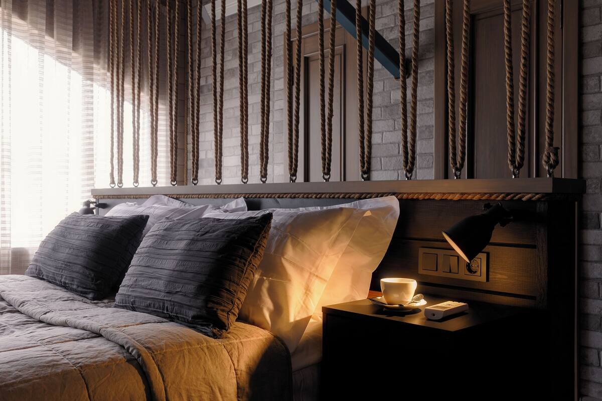 Un dormitorio con cama, almohadas, una mesita de noche con lámpara y una pared decorada con un diseño de cuerda.