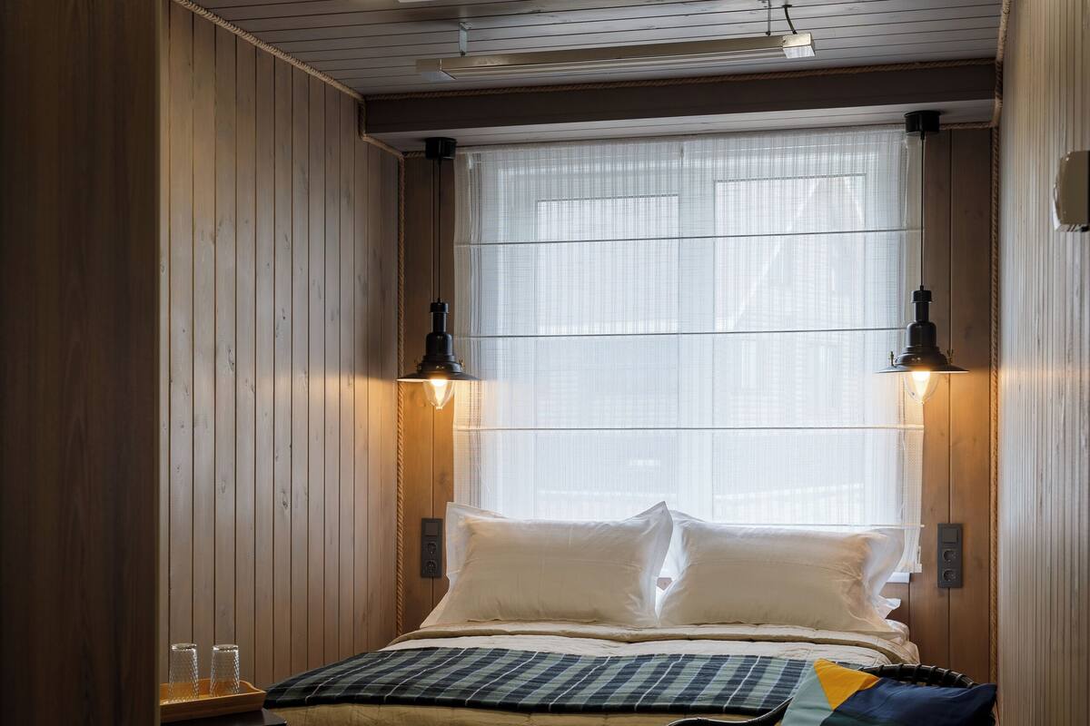 Un dormitorio con cama, lámparas de noche, una silla y una ventana con cortinas traslúcidas.