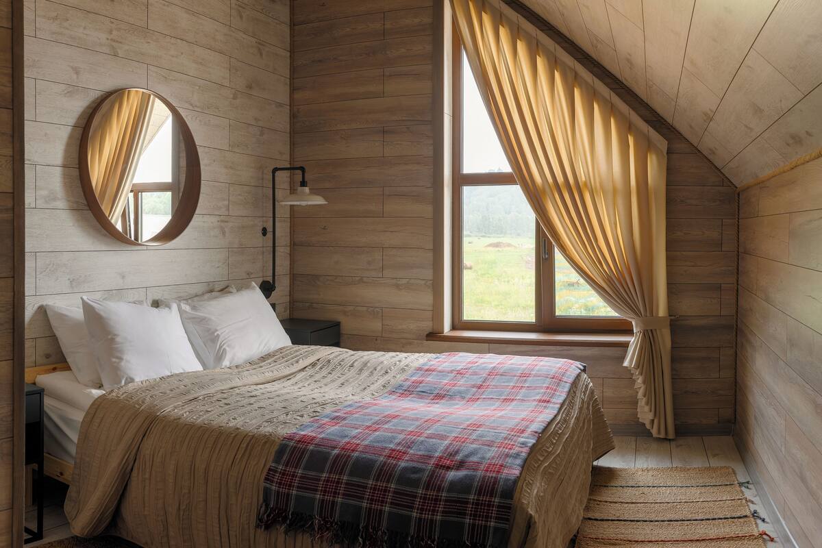 Un dormitorio con suelo de madera, una cama con manta escocesa, un espejo redondo, una mesita de noche, una lámpara y una ventana con cortinas.