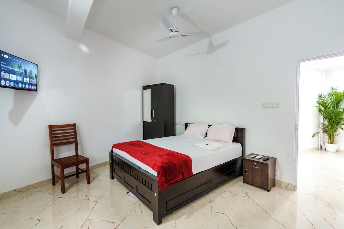 A bedroom with a bed, a chair, a wardrobe, a nightstand, and a TV on the wall.