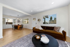 A spacious living area with a brown sofa, a dining table with chairs, and a kitchen in the background.