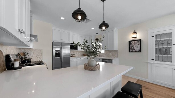 Une cuisine moderne avec des meubles blancs, des appareils en acier inoxydable, un grand îlot central, des luminaires suspendus et une plante en pot.