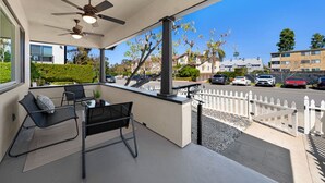Un espace extérieur aménagé, abrité, avec des chaises et une table, un ventilateur de plafond, et une vue sur une rue bordée de voitures garées et d’immeubles.