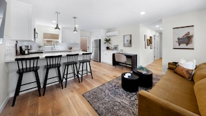 A modern kitchen with white cabinets, a central island with bar stools, a dining area with a black table and chairs, and a living room with a sofa and a coffee table.
