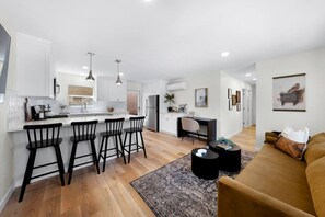 A modern kitchen with white cabinets, a central island with bar stools, a dining area with a black table and chairs, and a living room with a sofa and a coffee table.