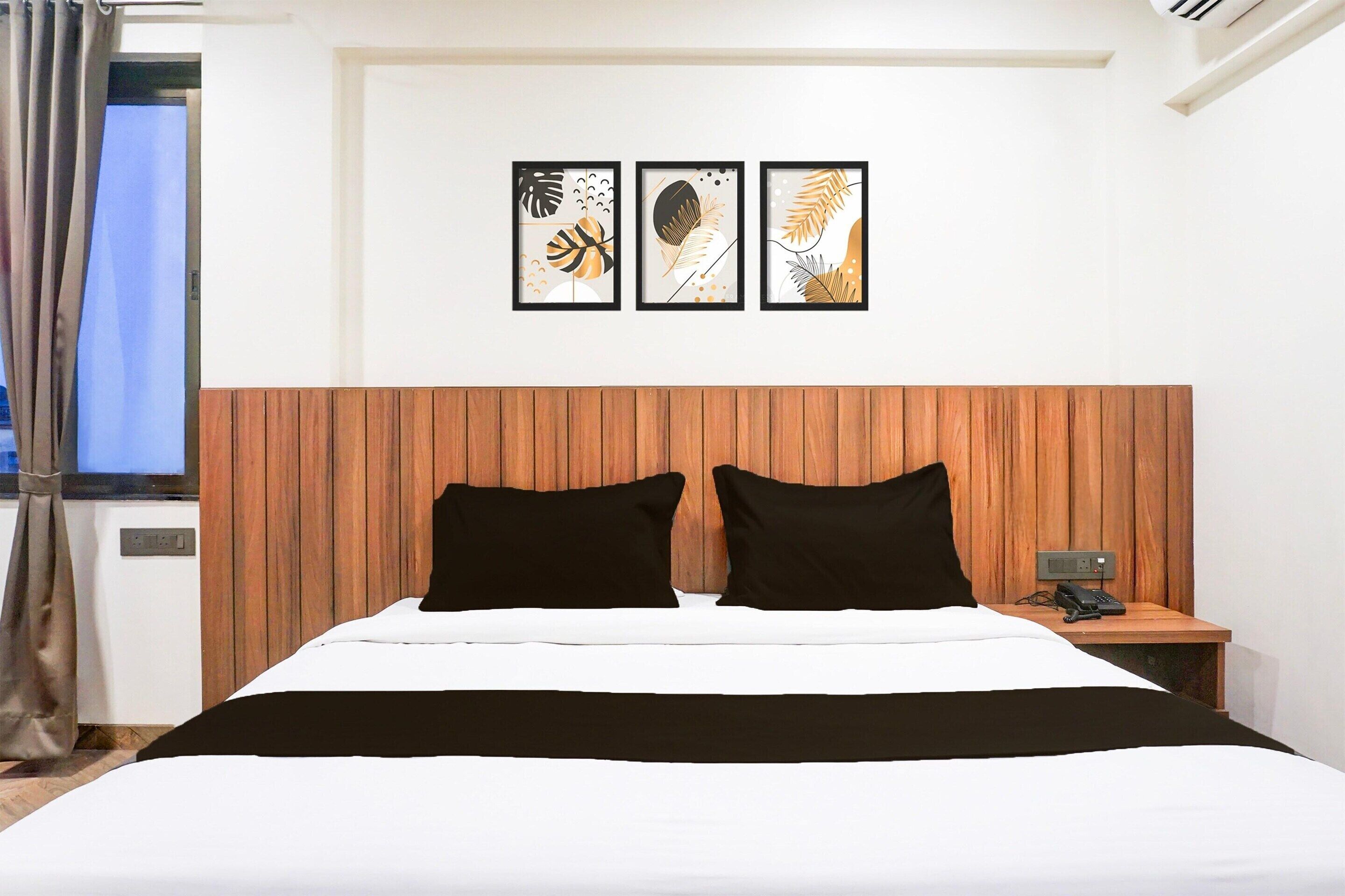 A hotel room with a wooden headboard, a bed with black and white bedding, a nightstand with a phone, and three framed wall art pieces above the bed.