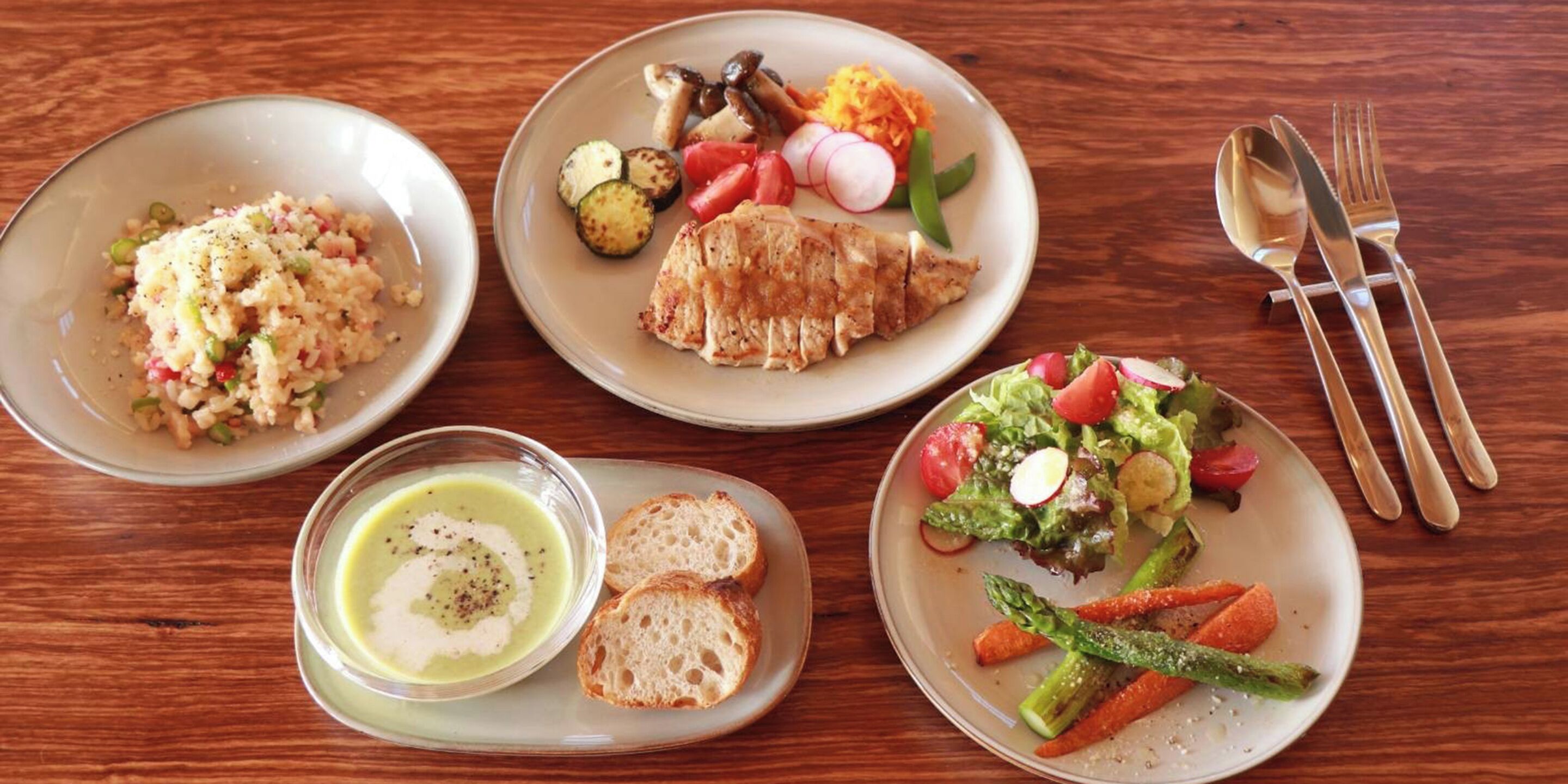 A table with four plates of food, including rice, grilled chicken, and vegetables, and a bowl of soup.