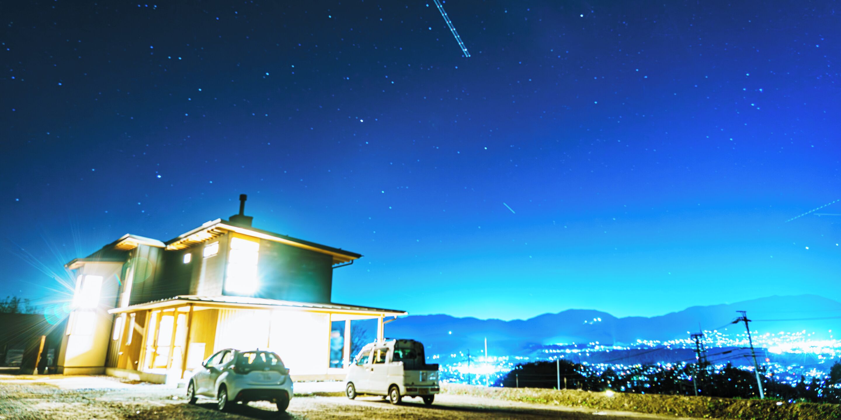 A house with a car parked in front, a truck parked to the side, and a clear night sky with stars.