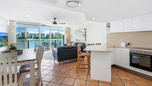 Una cocina moderna con armarios blancos, un comedor con mesa y sillas, un sofá negro y un amplio ventanal con vistas a una piscina y edificios.