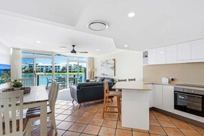 A modern kitchen with white cabinets, a dining area with a table and chairs, a black sofa, and a large window with a view of a pool and buildings.