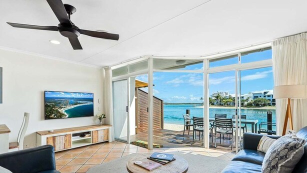 Una sala de estar moderna con televisor, ventilador de techo y vista a una playa y al océano a través de amplias puertas de cristal.