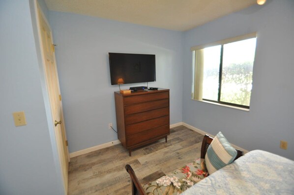 A bedroom with a wooden dresser, a television on the wall, a window with a view, and a bed with a floral bedspread.