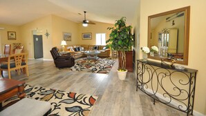 A spacious living room with a high ceiling, a large plant, a dining area with wooden furniture, and a console table with a mirror and decorative items. - Salty Breeze Canal Front Pool Home W Boat Dock (Palm Coast)