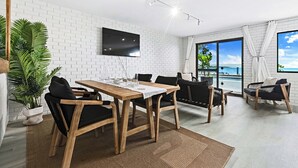 A modern living room with a wooden dining table, black cushioned chairs, a flat-screen TV mounted on a white brick wall, and a large window offering a view of the ocean.