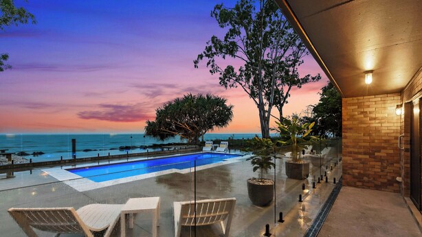 A rooftop pool with lounge chairs, a glass balustrade, and a view of the ocean at sunset.