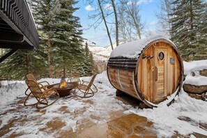 A wooden barrel sauna surrounded by snow-covered ground, with a fire pit and wicker chairs nearby.