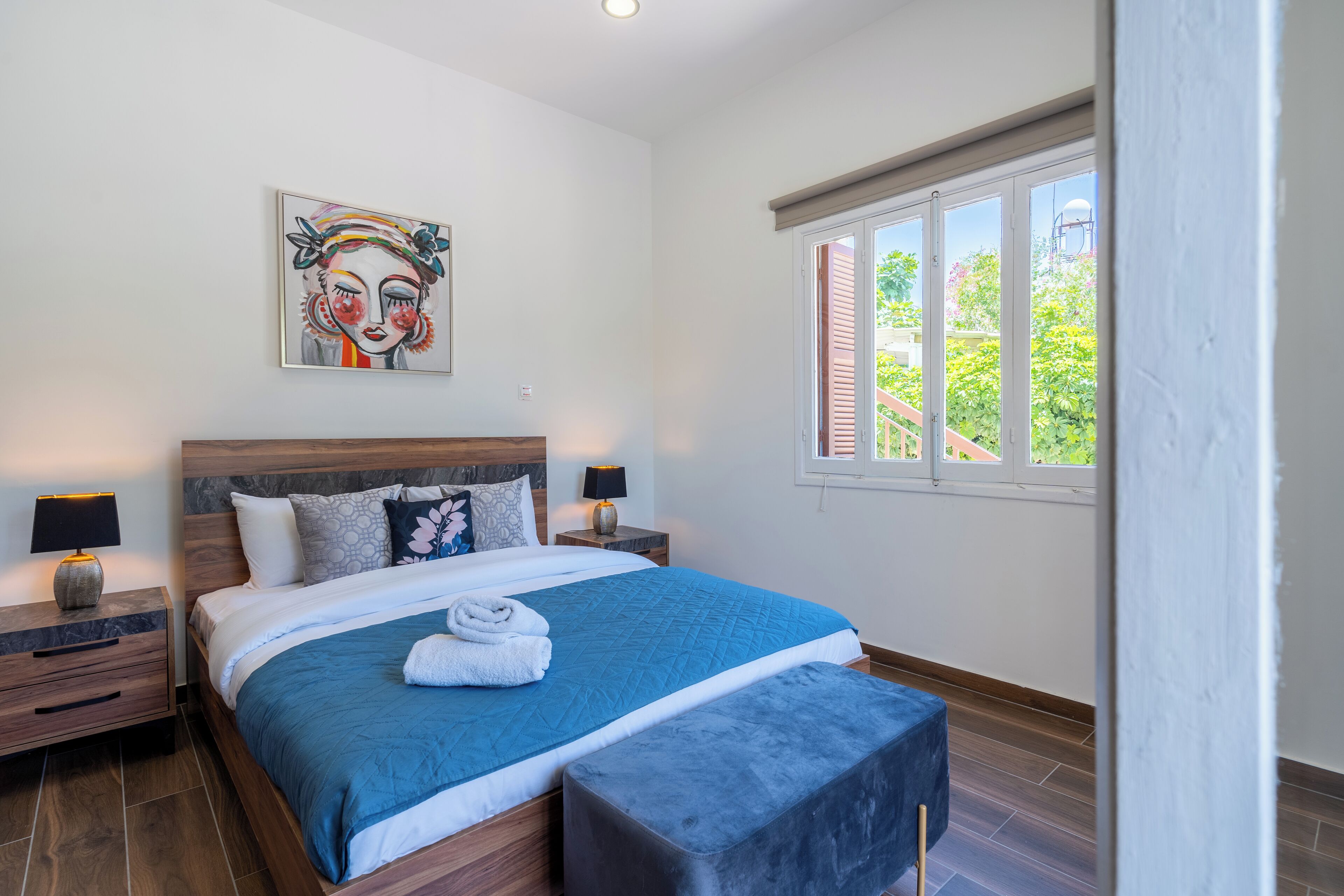 Une chambre avec un grand lit, deux tables de chevet avec des lampes, un tableau au mur et une fenêtre avec des volets.