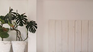 Un dormitorio con cabecera de madera, una cama blanca, una mesita redonda con una cesta y dos plantas grandes en macetas.