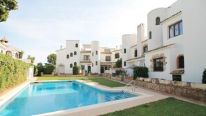 Zona de piscina con una piscina azul cristalina, rodeada de edificios blancos con ventanas de arco y muros de piedra.
