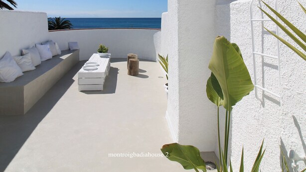 Una terraza en la azotea con un muro blanco, una zona de estar pequeña con cojines y vistas al mar.