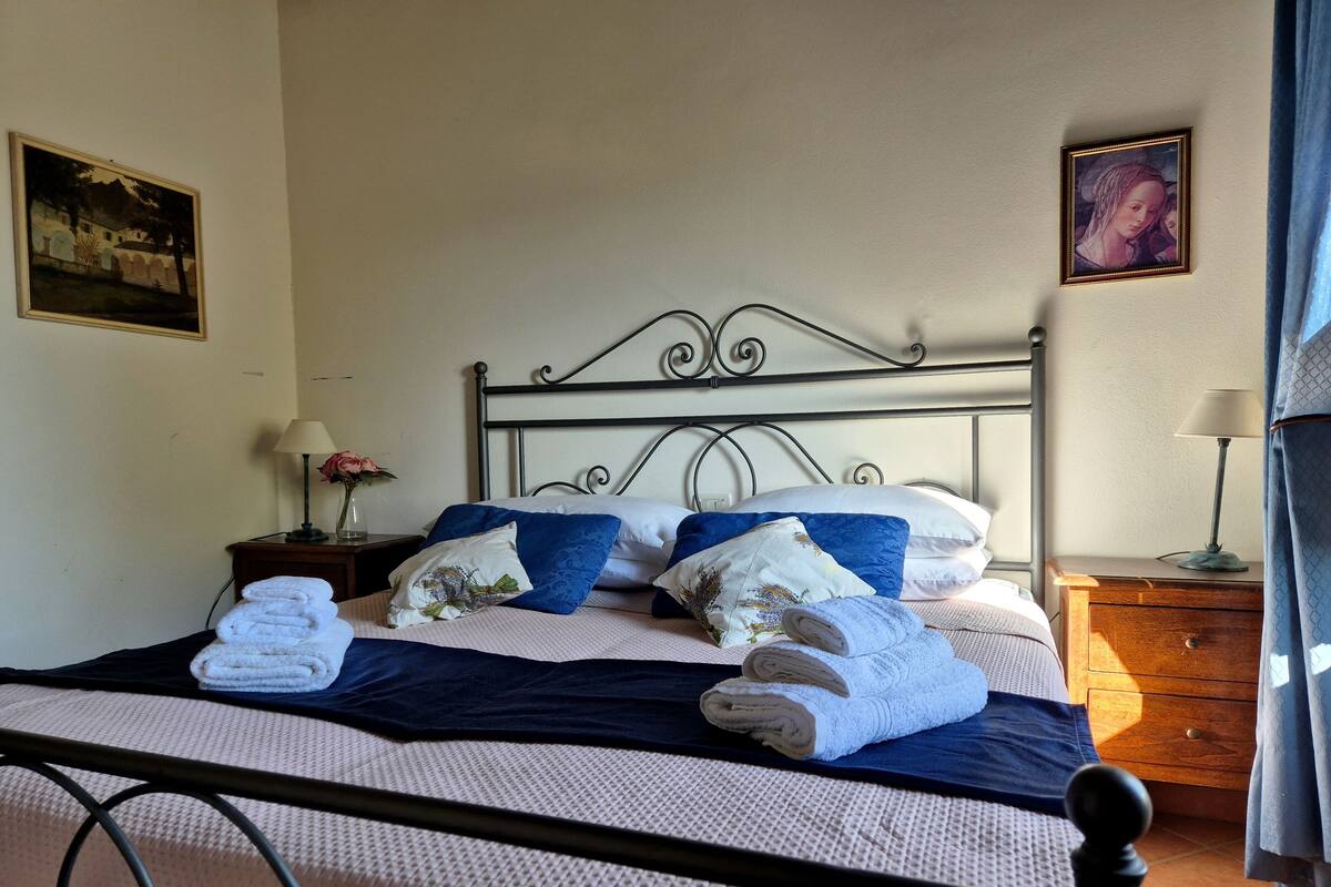 Een net opgemaakt bed met kussens en opgevouwen handdoeken, een nachtkastje met een lamp, en een schilderij aan de muur.