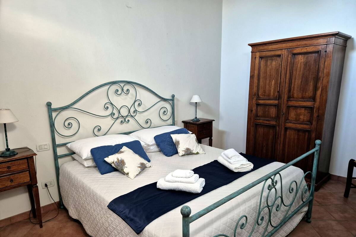 Een net opgemaakt bed met een gewatteerde sprei, twee kussens en opgevouwen handdoeken. Er staat een houten garderobe en een nachtkastje met een lamp naast het bed.