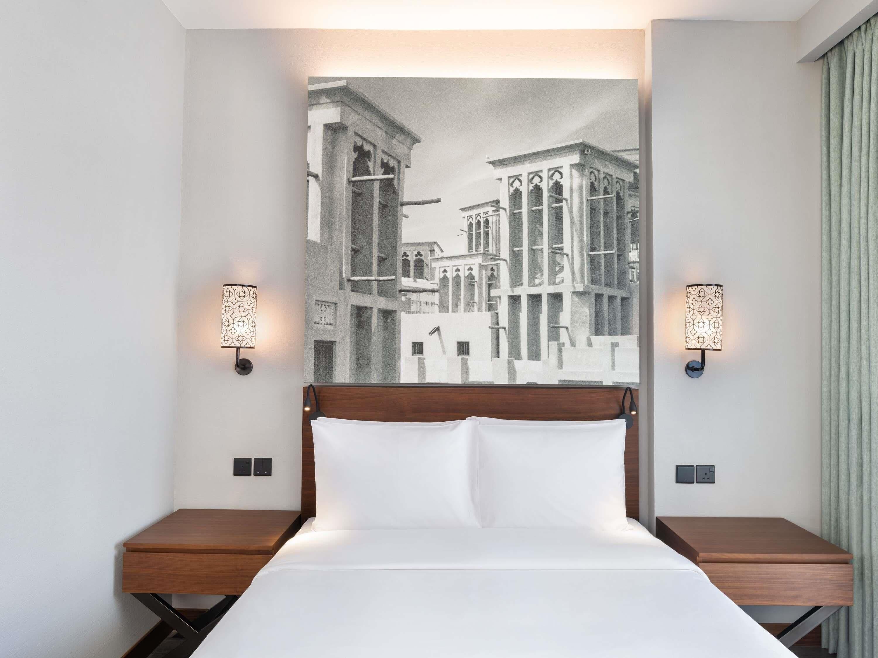 Habitación de hotel con una cama, dos mesitas de noche, un cabecero y una obra de arte colgada en la pared que representa un paisaje urbano.