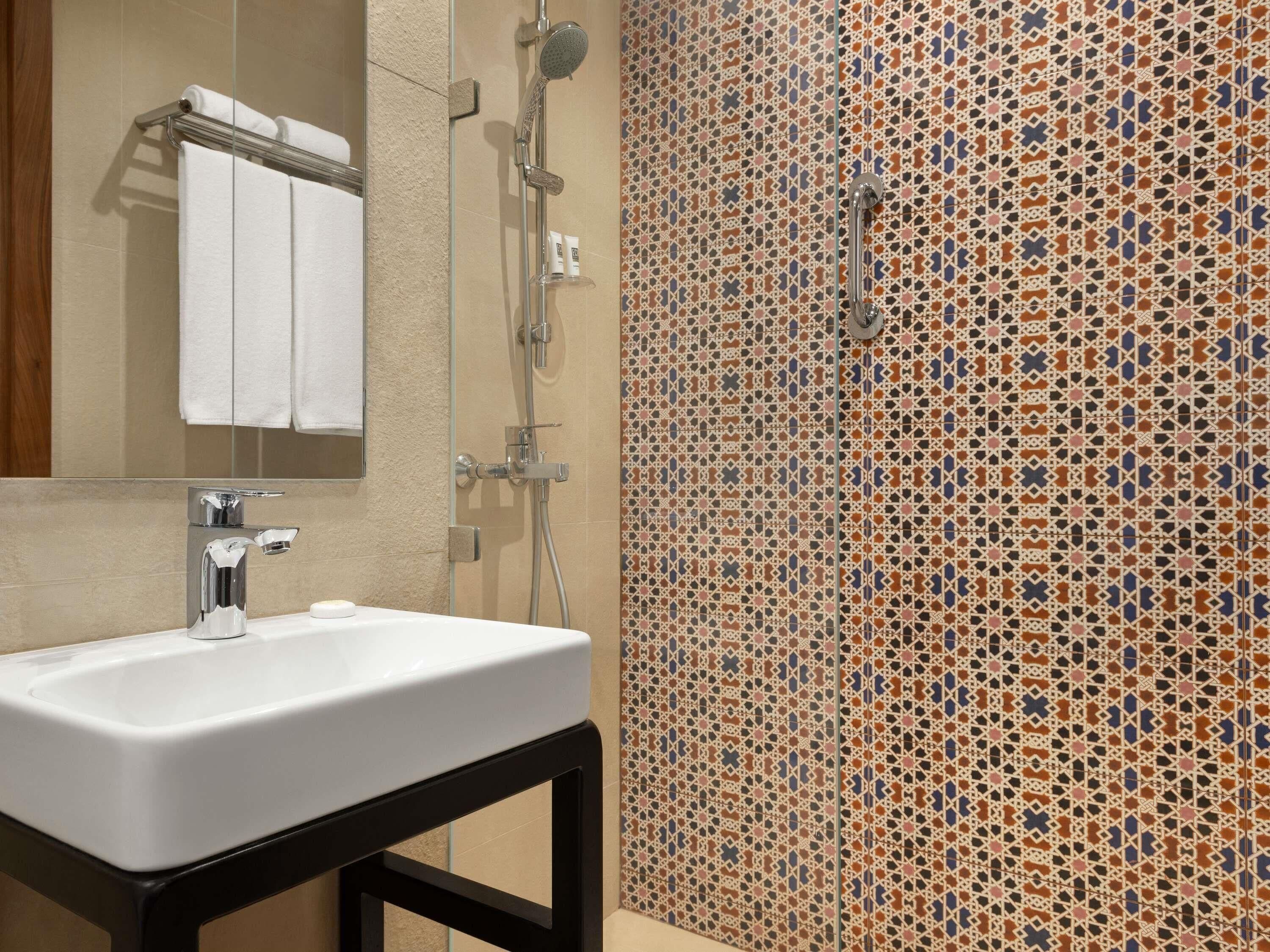 A bathroom with a white sink, a mirror, a shower with a handheld showerhead, and a tiled wall with a geometric pattern.