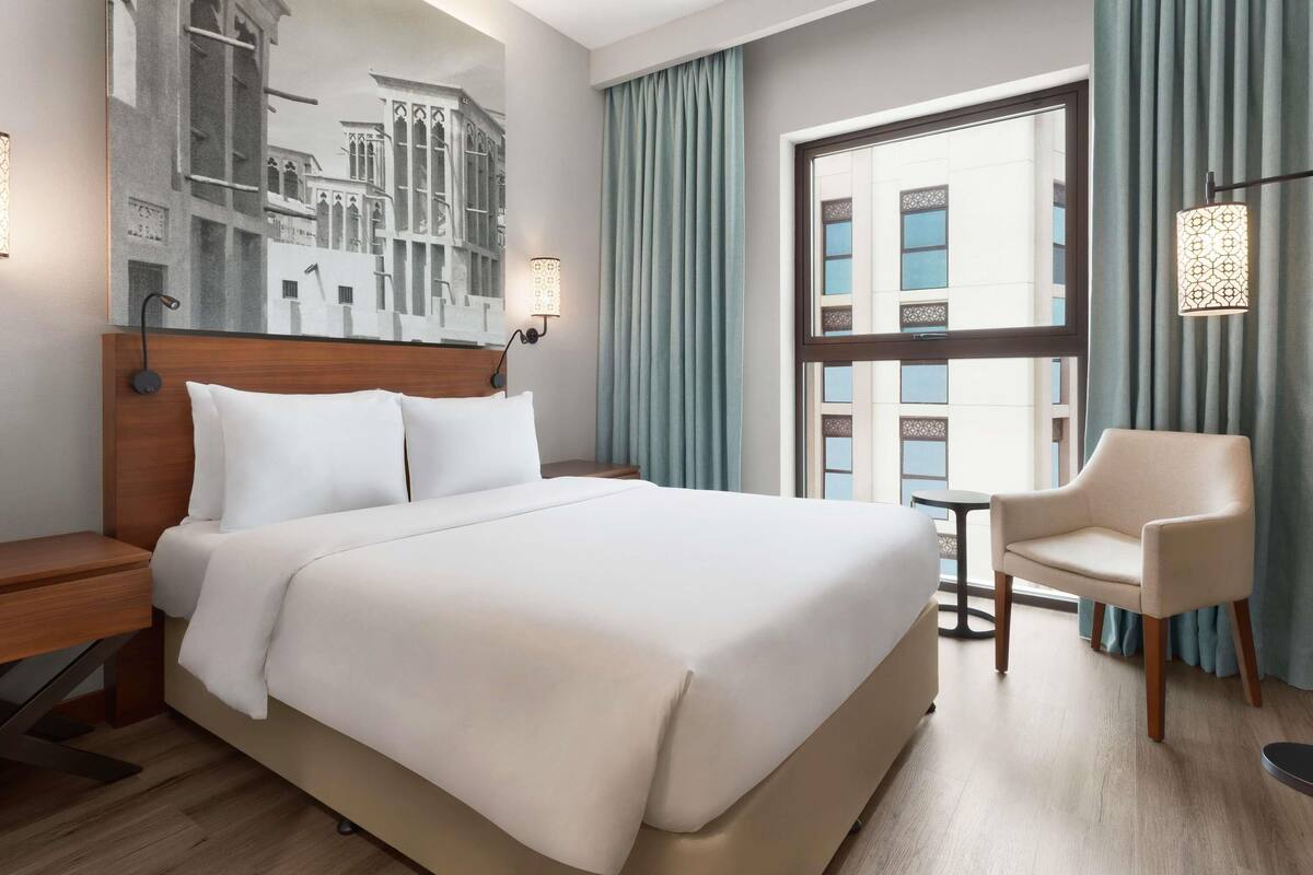 Ein Hotelzimmer mit einem großen Bett, einem Nachttisch, einem Sessel, einem kleinen Tisch, einem Fenster mit Vorhängen und einer Wandlampe.