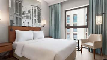 Une chambre d’hôtel avec un grand lit, une table de chevet, une chaise, une petite table, une fenêtre avec des rideaux et une lampe fixée au mur.