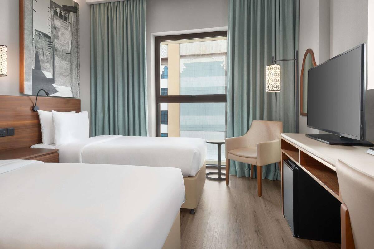 Ein Hotelzimmer mit zwei Betten, einem Schreibtisch, einem Fernseher und einem Fenster mit Vorhängen.