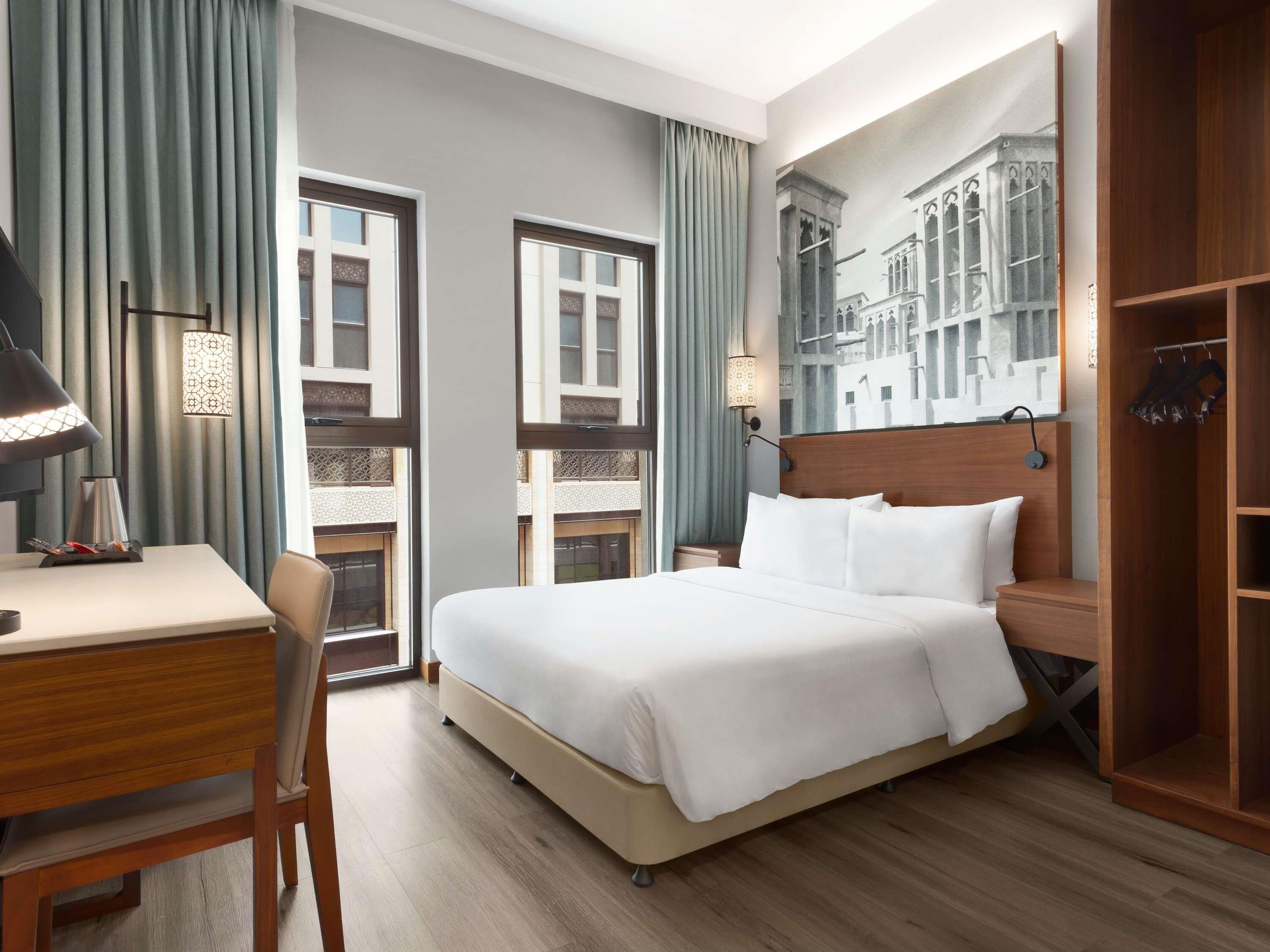 Une chambre d’hôtel avec un grand lit, un bureau, une chaise et une armoire. Elle comprend deux fenêtres avec des rideaux, une lampe fixée au mur et un grand miroir.