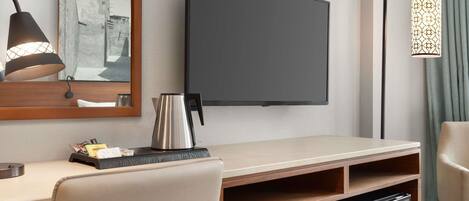 A hotel room with a flat-screen TV mounted on the wall, a desk with a kettle, a chair, and a small refrigerator.