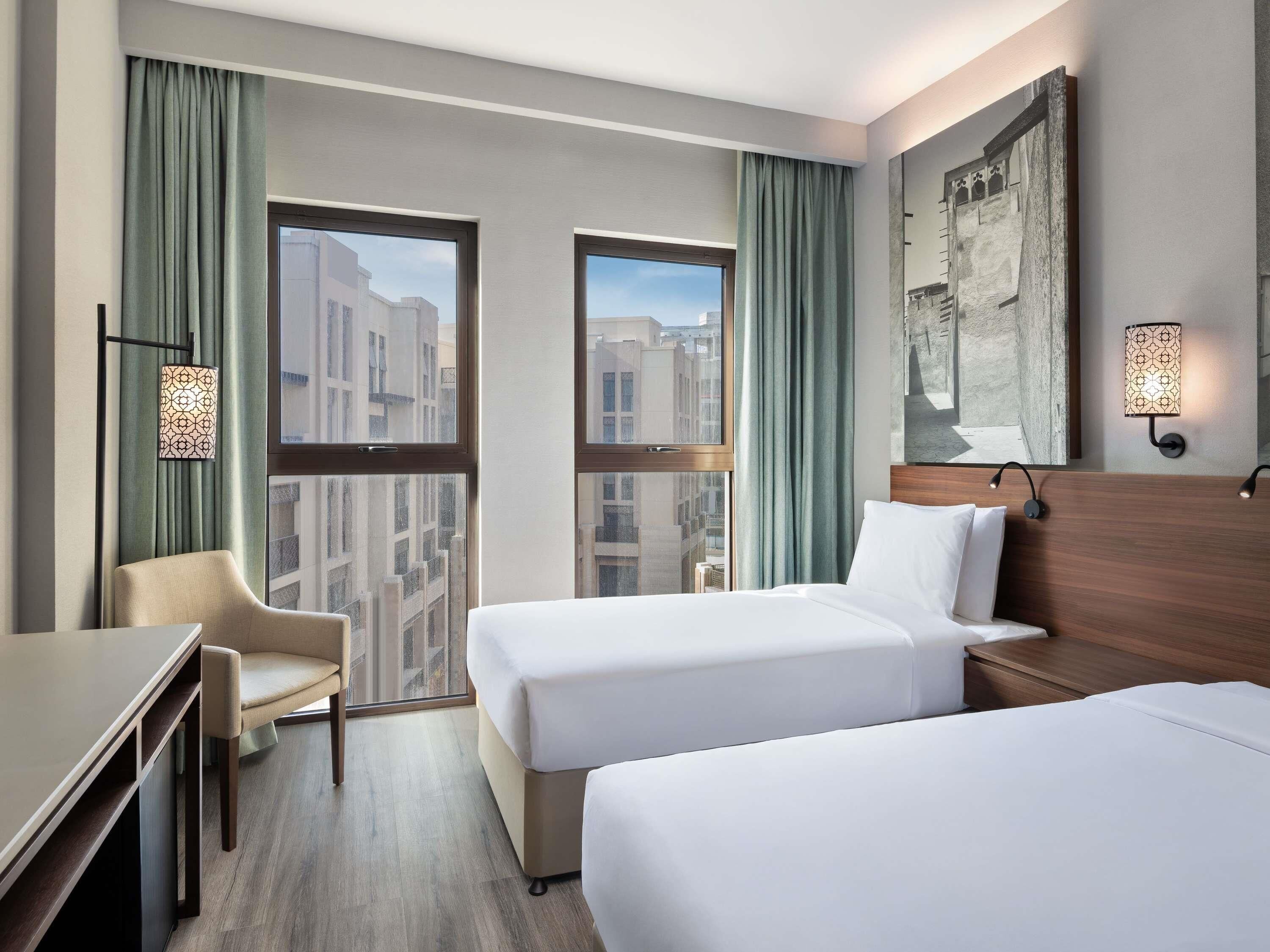 Habitación de hotel con dos camas, un escritorio, una silla y ventanas amplias con cortinas.
