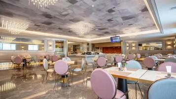 Een ruime hotellobby met modern interieur, een receptie en een eetgedeelte met ronde tafels en stoelen.