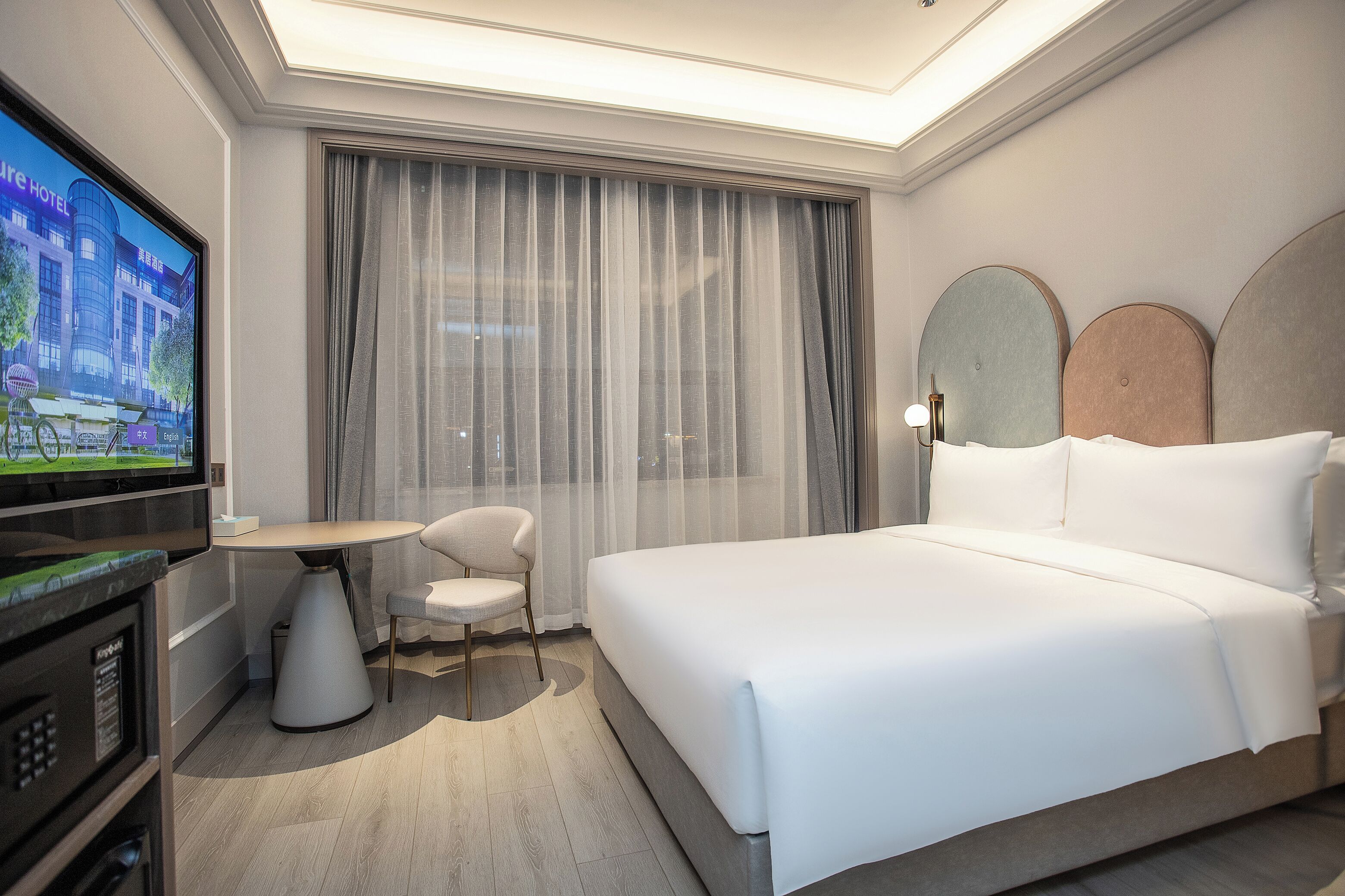 A hotel room with a large bed, a television, a small table, a chair, and a window with curtains.