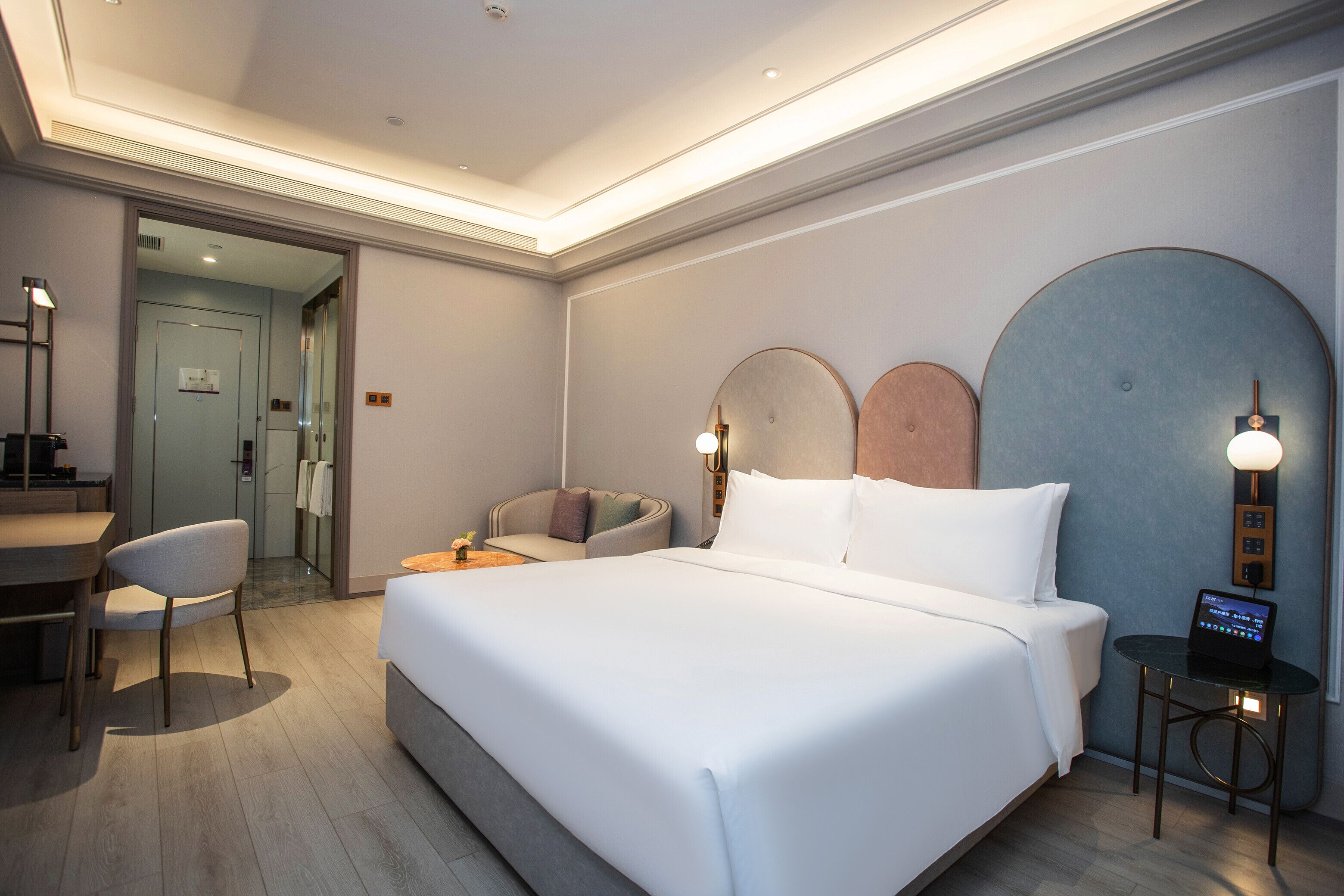 A modern hotel room with a large bed, a desk, a chair, and a small table with a laptop.