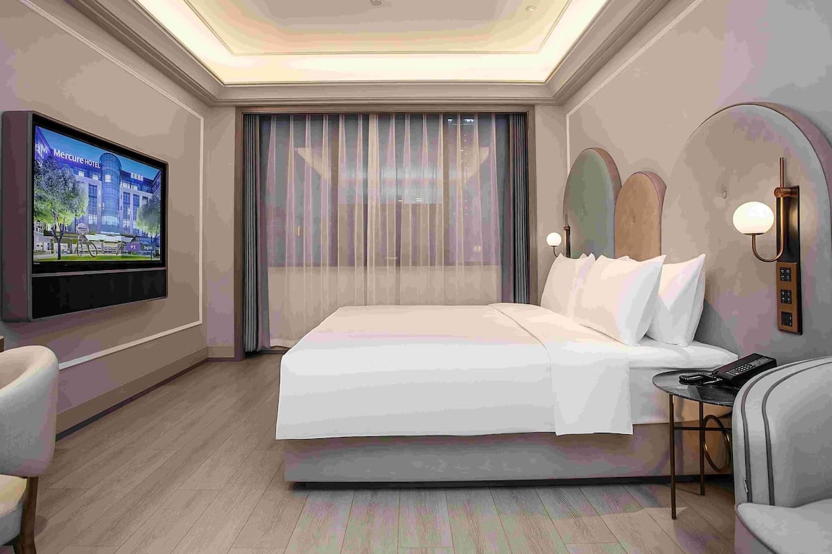 A modern hotel room with a large bed, a television mounted on the wall, a desk with a phone, and a chair.