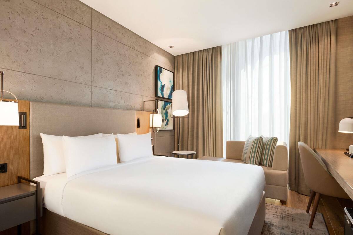 A modern hotel room with a large bed, a desk, a chair, and a view of the outdoors through large windows.