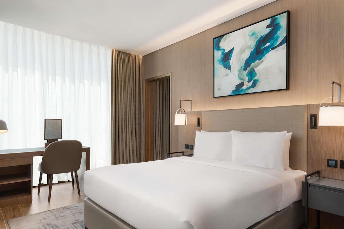 A modern hotel room with a large bed, a desk with a chair, a desk lamp, and a painting on the wall.