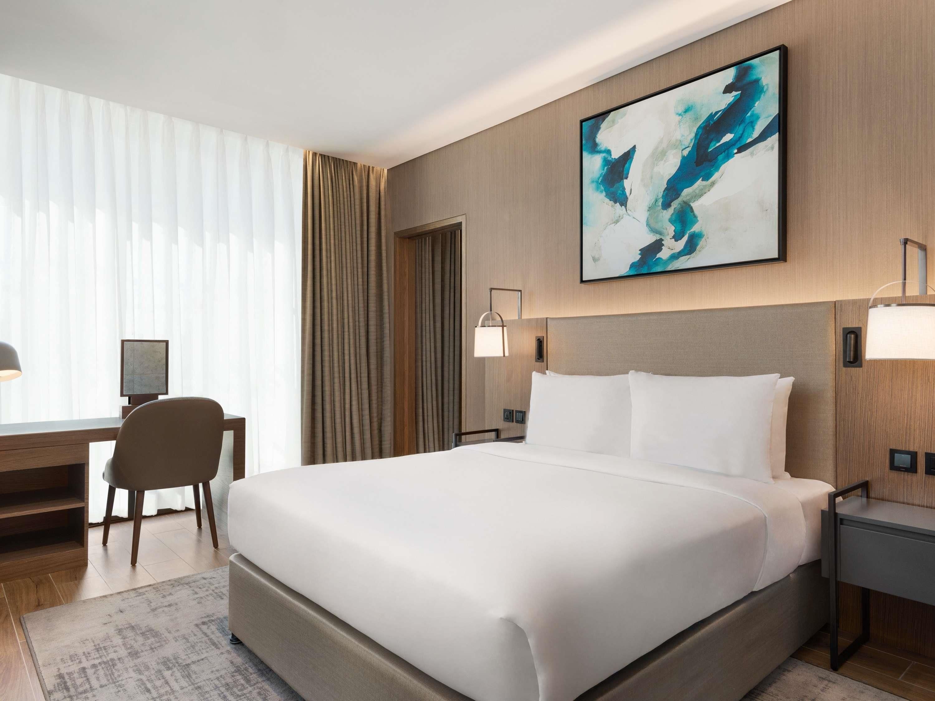 Une chambre d’hôtel moderne avec un grand lit, un bureau avec une chaise, une lampe de bureau et un tableau au mur.