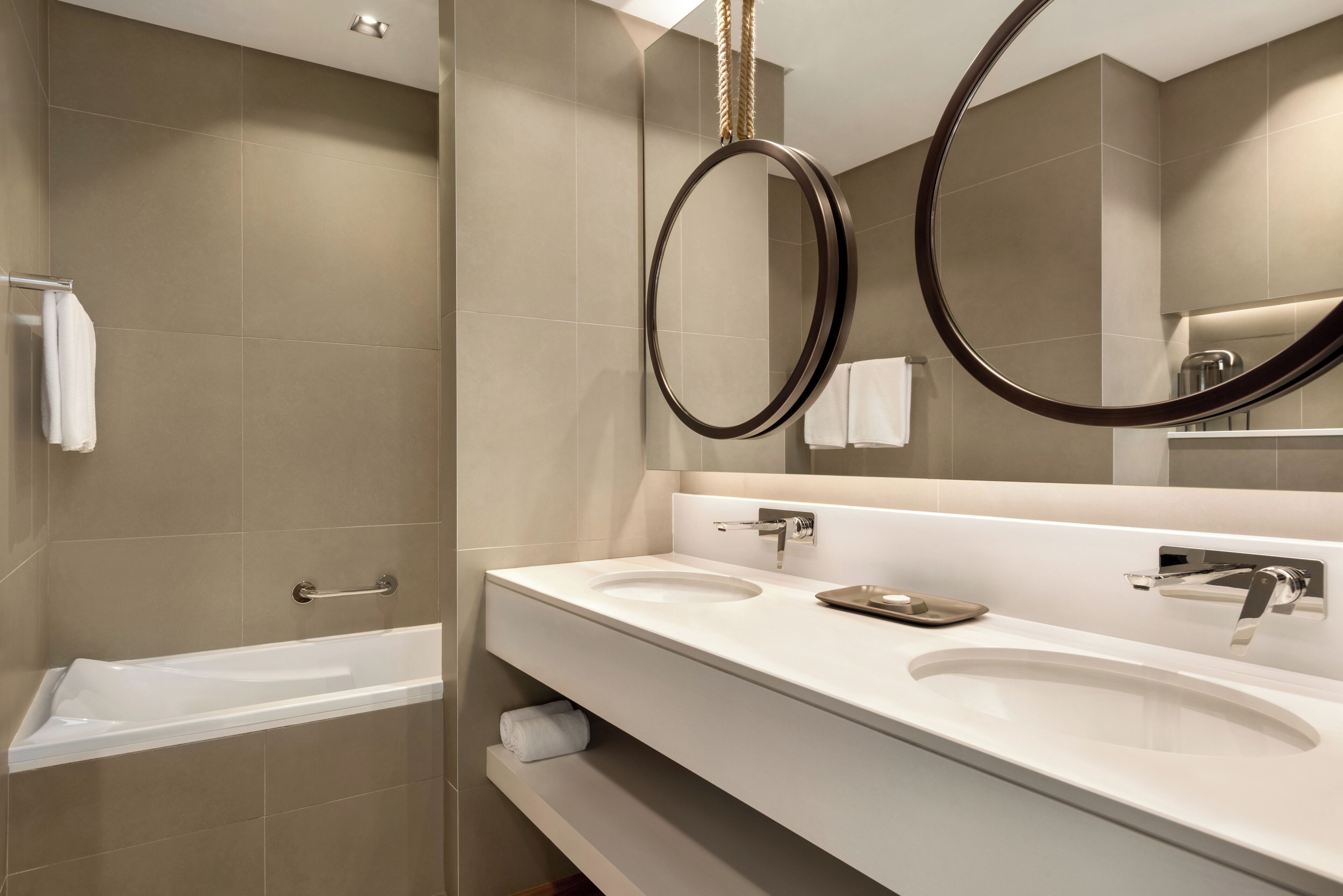 A modern bathroom with a bathtub, two sinks, and a large mirror with a circular frame.