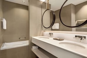 A modern bathroom with a bathtub, two sinks, and a large mirror with a circular frame. - Mercure Dubai Gold District (Dubai)