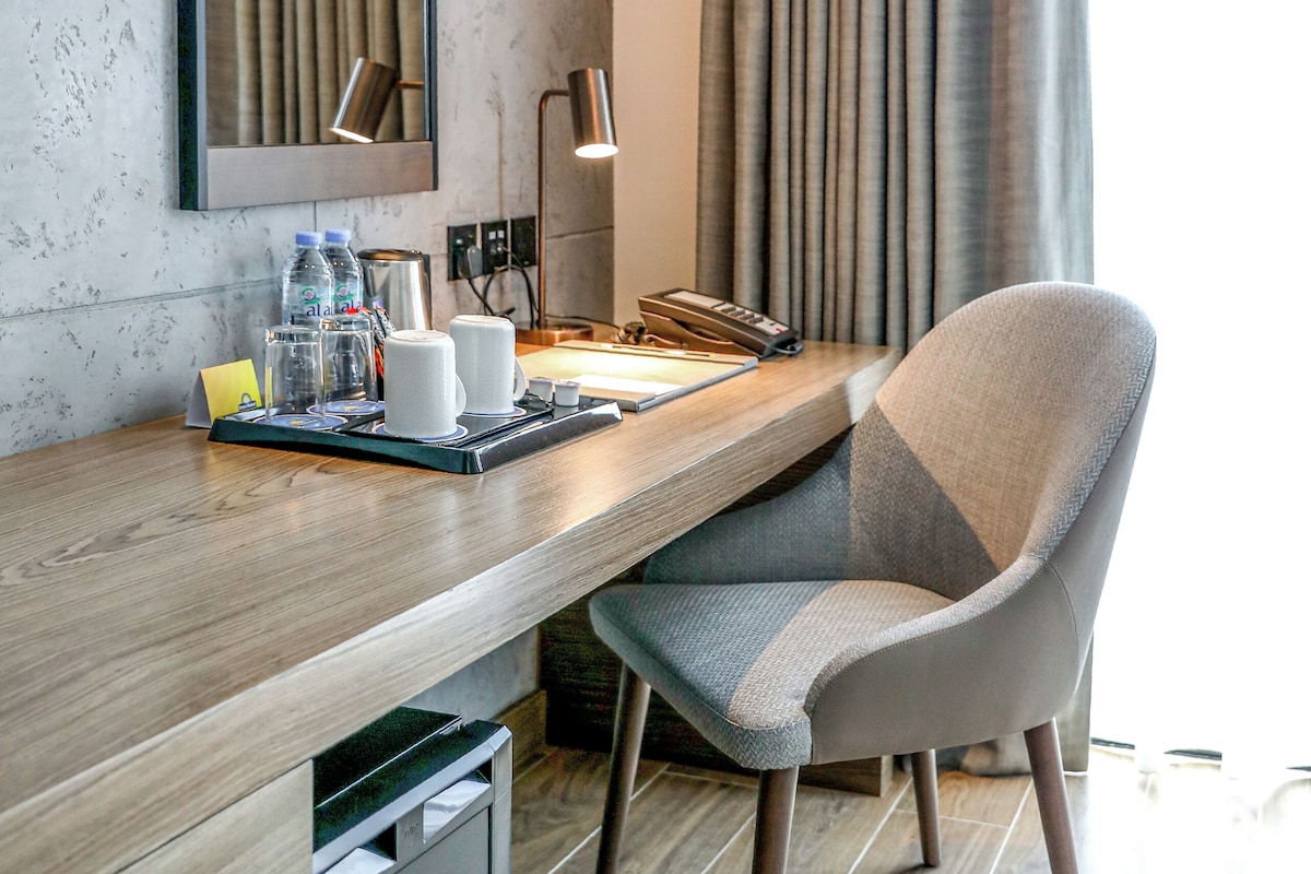 A modern hotel room with a wooden desk, a chair, a mirror, and a view of the outside through curtains.