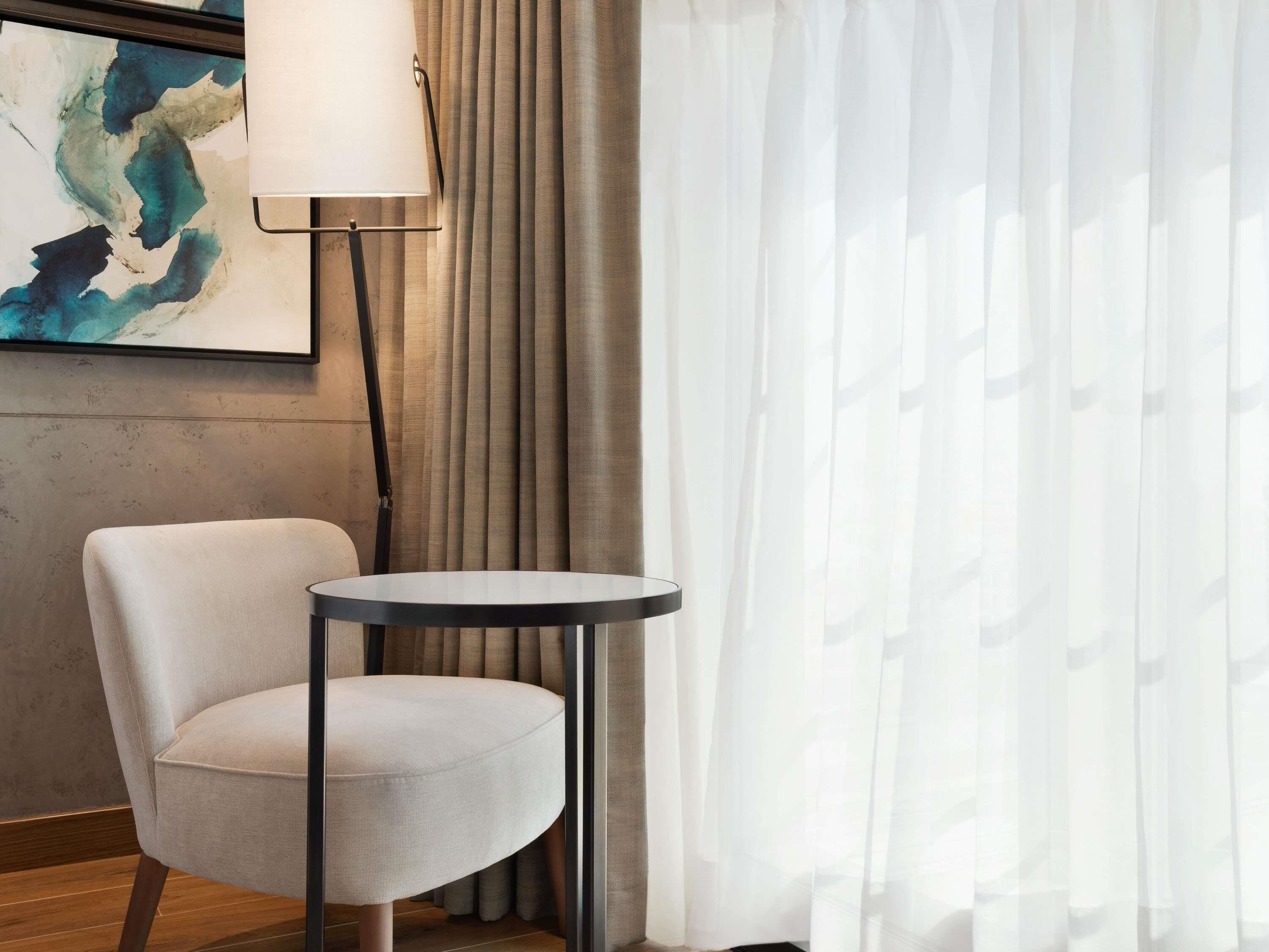 A room with a round table, a chair, a lamp, and a painting on the wall.