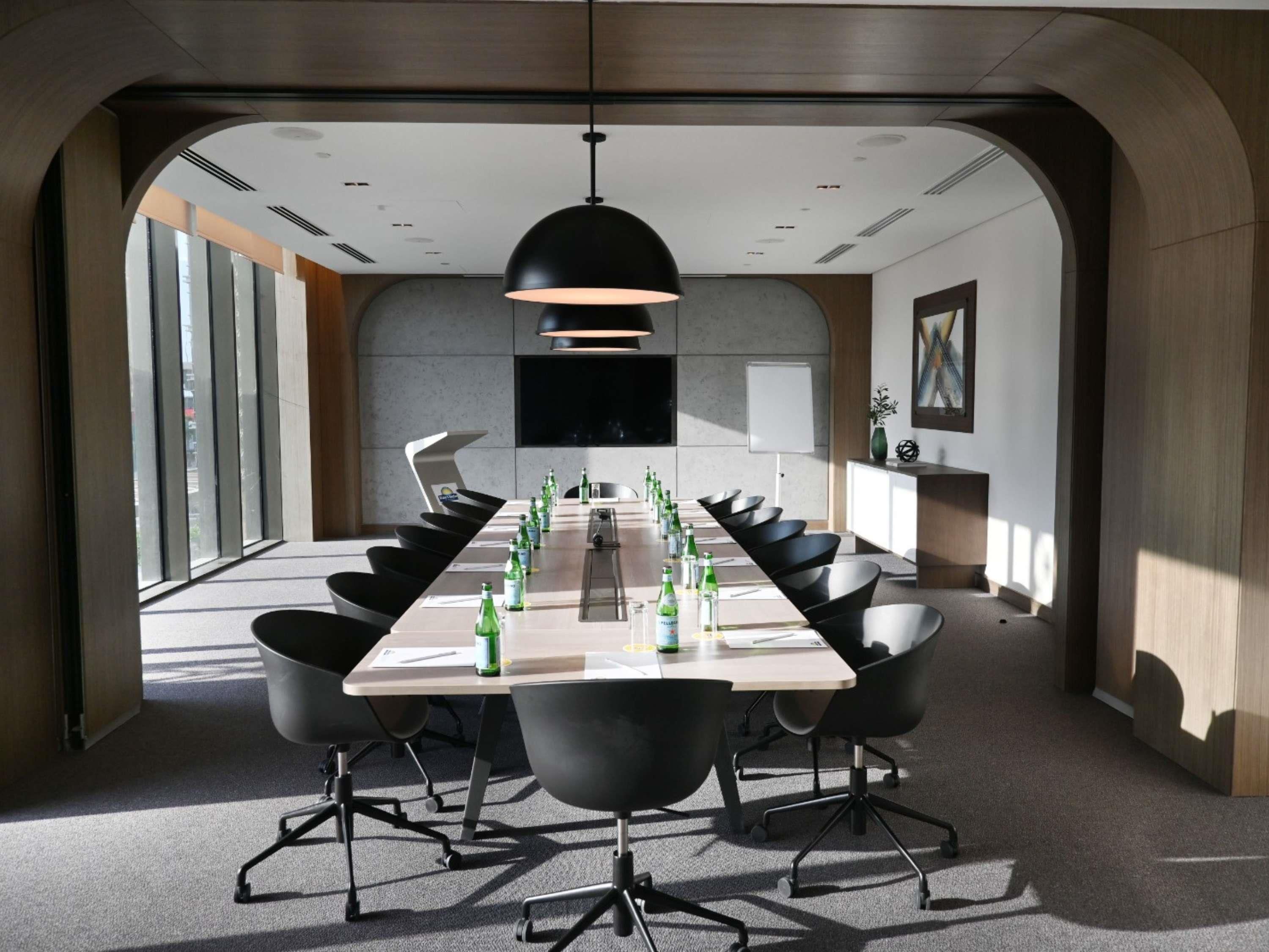 Ein Konferenzraum mit einem langen Tisch, schwarzen Stühlen, einer schwarzen Pendelleuchte und einer Whiteboard-Tafel.
