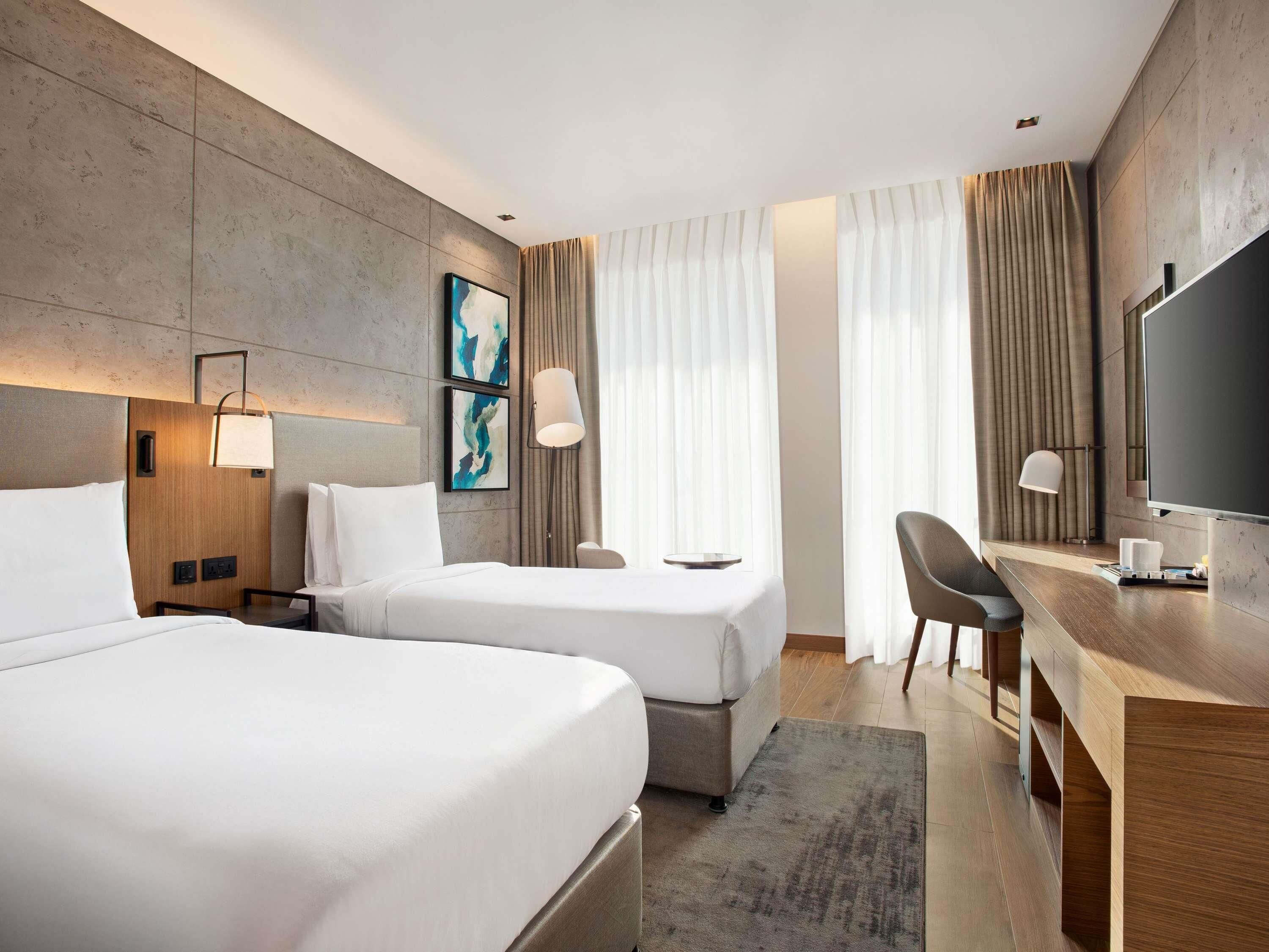 Ein modernes Hotelzimmer mit zwei Betten, einem Schreibtisch, einem Sessel, einem Fernseher und Kunstwerken an den Wänden.